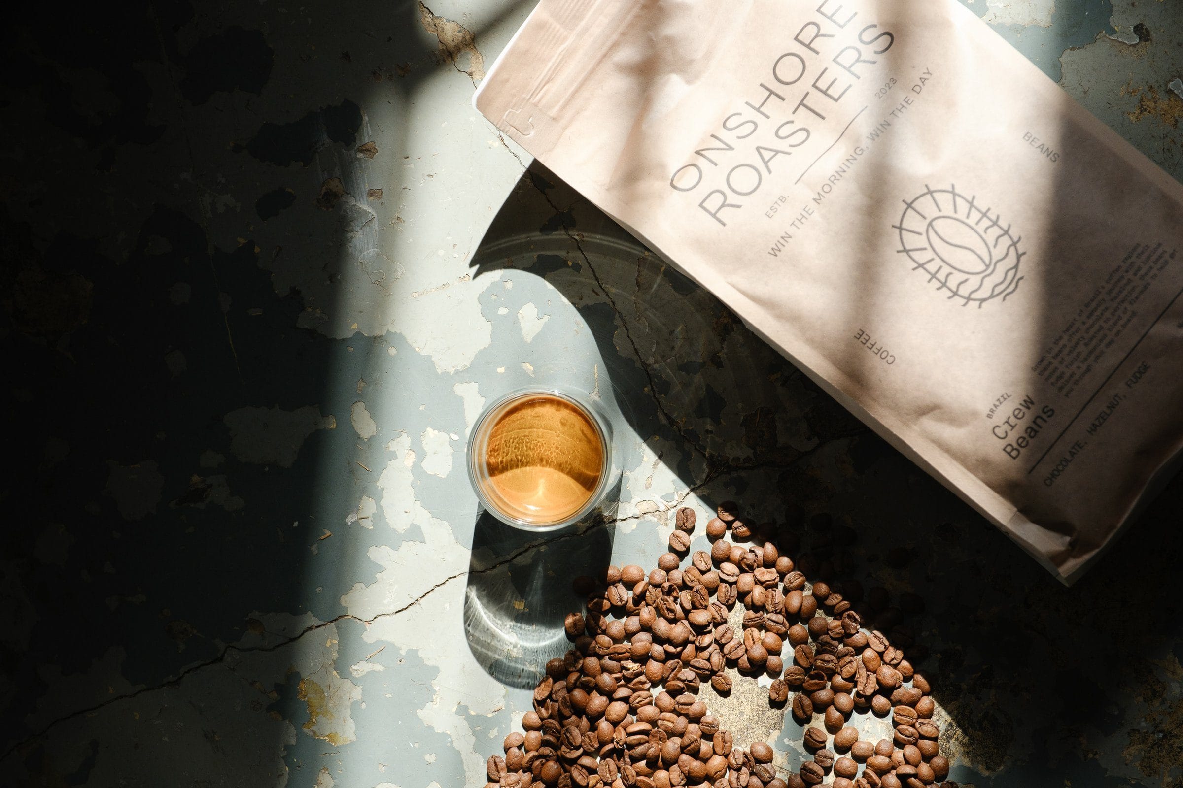 Una bolsa de Crew Beans, un espresso en vaso y granos de café tostados