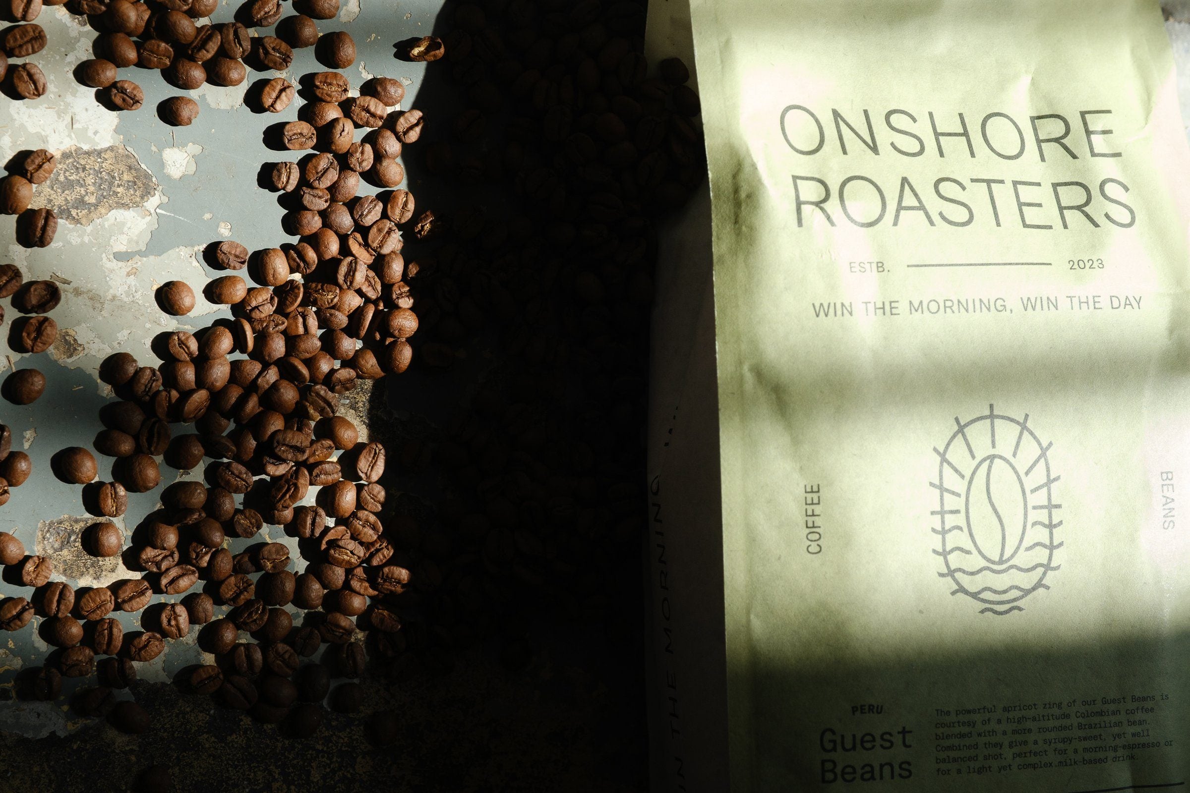 Granos de café tostados esparcidos junto a una bolsa de Guest Beans a pleno sol